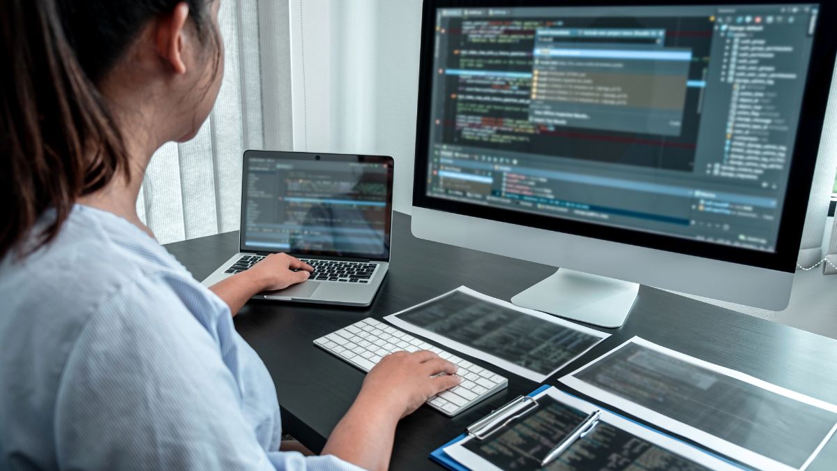 Egy programozó laptopon és asztali számítógépen kódot ír, papírok mellett.