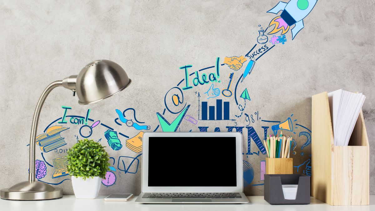 Egy modern íróasztal laptopja, mellette lámpa és növény, inspiráló grafikai elemekkel a falon.