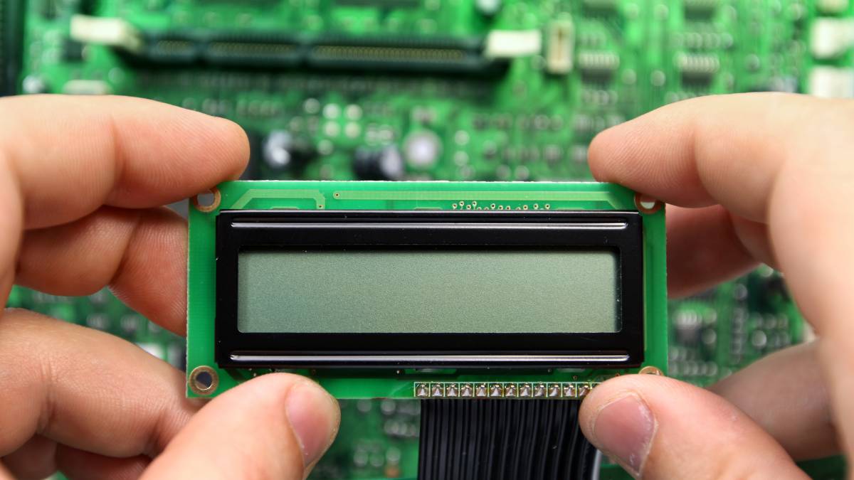 Kézben tartott LCD kijelző, háttérben elektronikai áramkör látható.